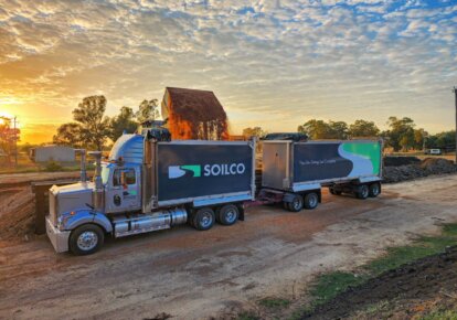 Yellow excavator loading soil into a white truck with the SOILCO logo on it. Sunset sky