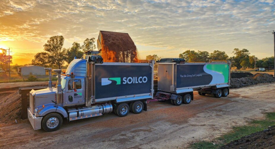 Yellow excavator loading soil into a white truck with the SOILCO logo on it. Sunset sky